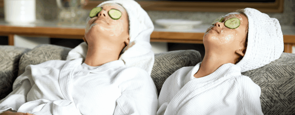 Two people enjoy a home spa, wearing white robes and towels on their heads as they relax on a couch with facial masks and cucumber slices over their eyes.
