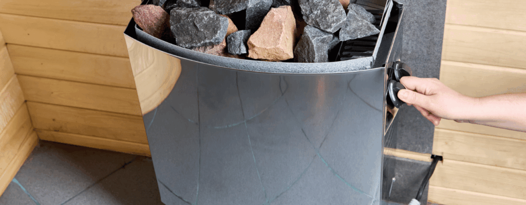 A person grips a large metal bin containing rocks, intended for an electric sauna setup.