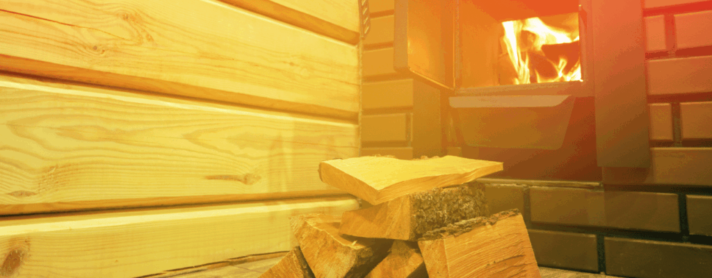 Stacked firewood sits on the floor near a lit wood-burning stove, evoking the cozy ambiance of a traditional wood fired sauna, with wooden paneling and brick walls in the background.