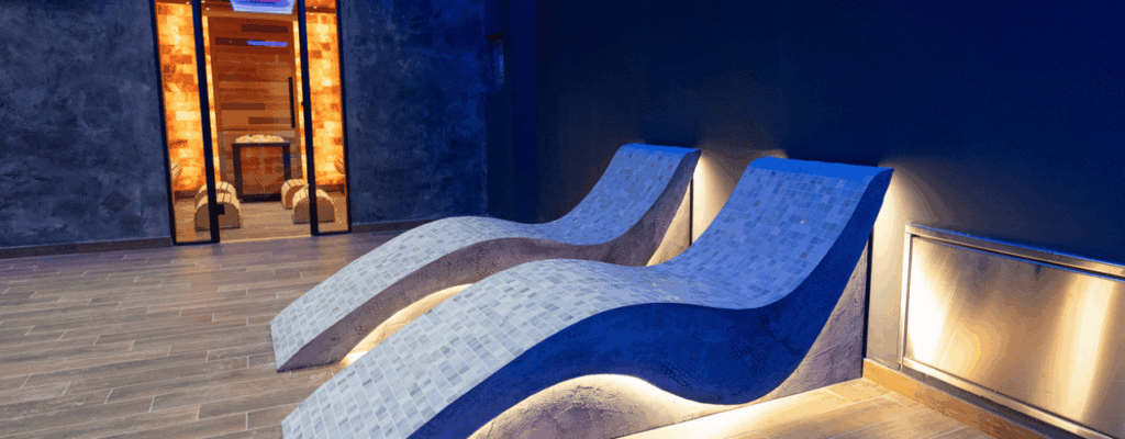 Modern wellness space showcasing elegant heated loungers and sauna entrance, illuminated by soothing ambient blue lighting, evoking calm and relaxation.