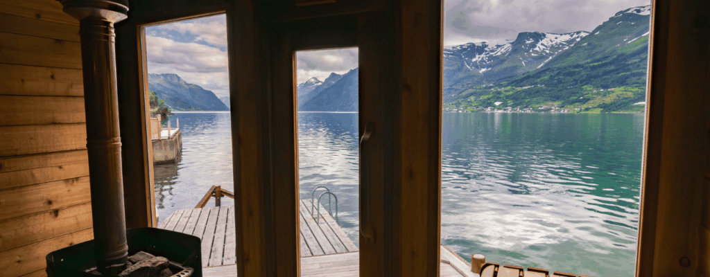 View from a wooden cabin showing a lake and mountains, with an outdoor sauna enhancing the tranquil setting.