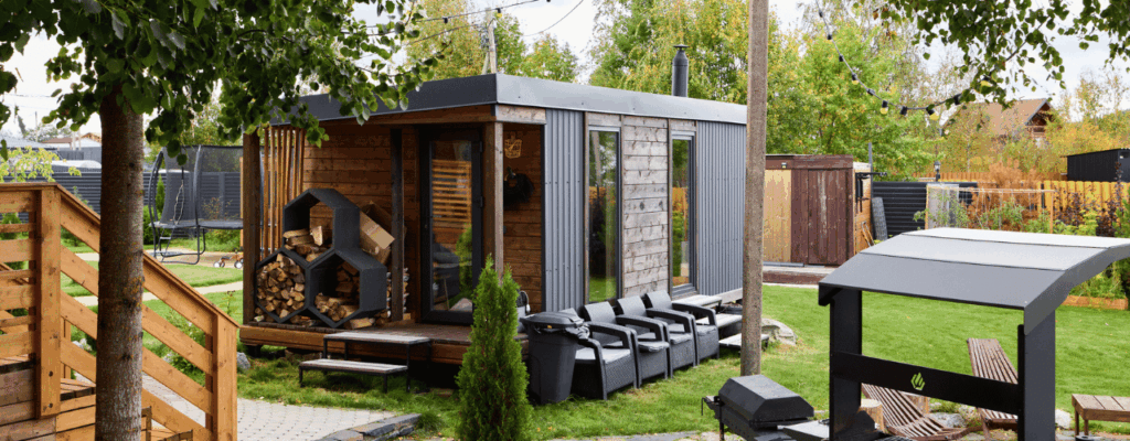Wooden backyard sauna building with stacked lounge chairs, cedar siding and outdoor smoker, manicured lawn and stone pathway